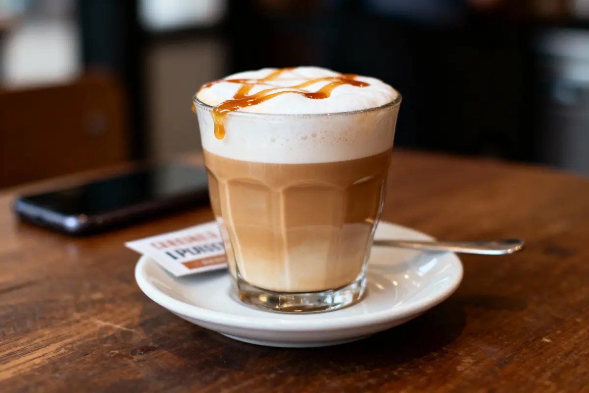Un café latte dans un verre transparent, avec une mousse de lait et un filet de caramel, posé sur une soucoupe.