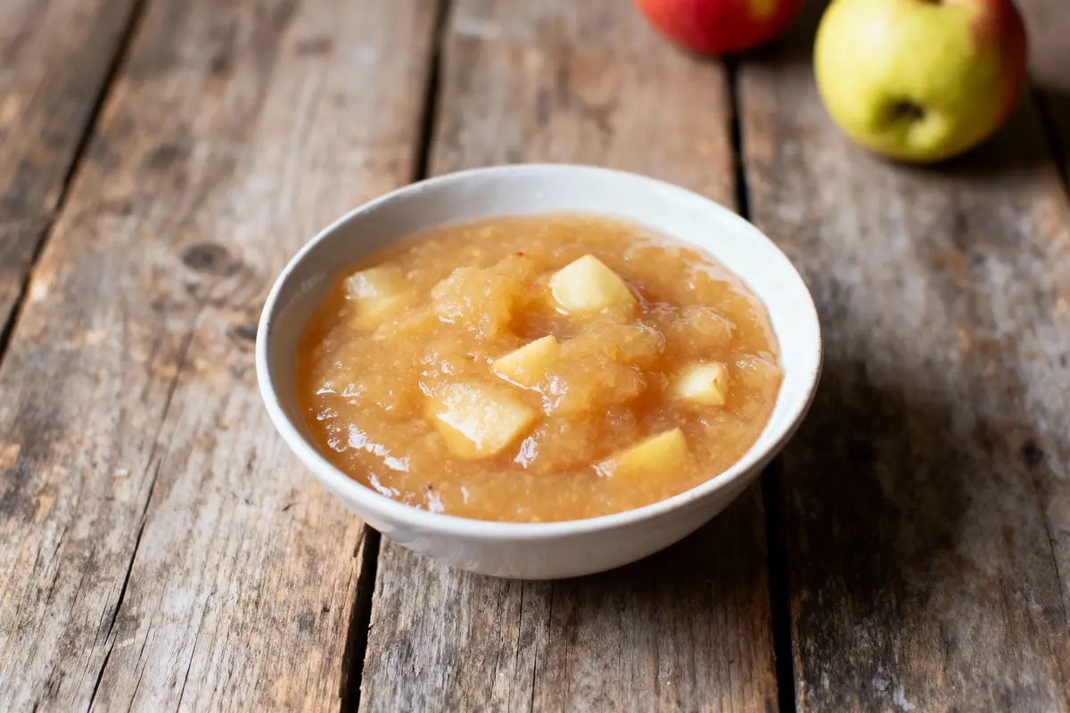 Bol de compote de pommes avec morceaux, sur une table en bois rustique, entouré de pommes entières.