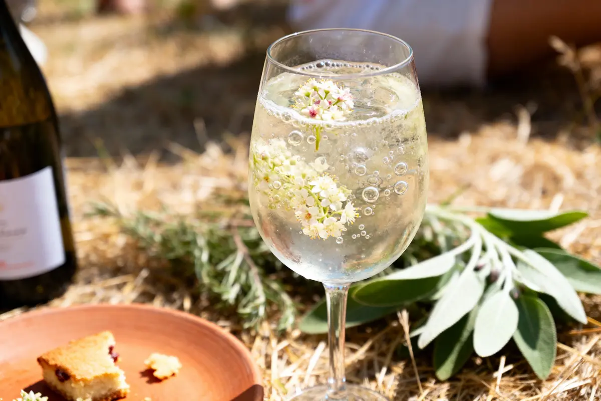 Un verre de boisson pétillante avec des fleurs, posé sur un sol en herbe sèche, à côté d'un plat et d'une bouteille.