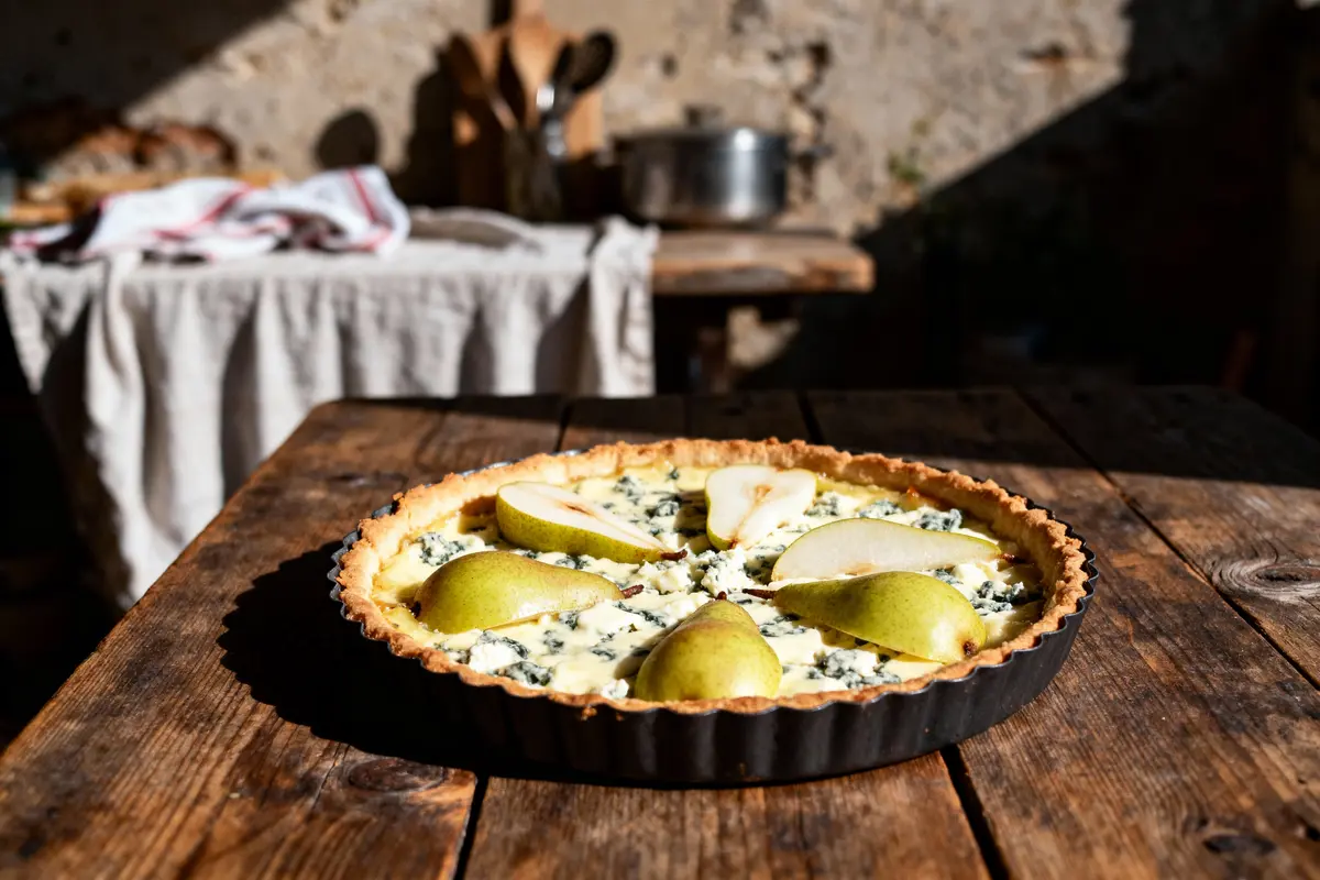Tarte aux poires sur une table en bois, garnie de fromage bleu, avec un fond naturel et un éclairage chaleureux.
