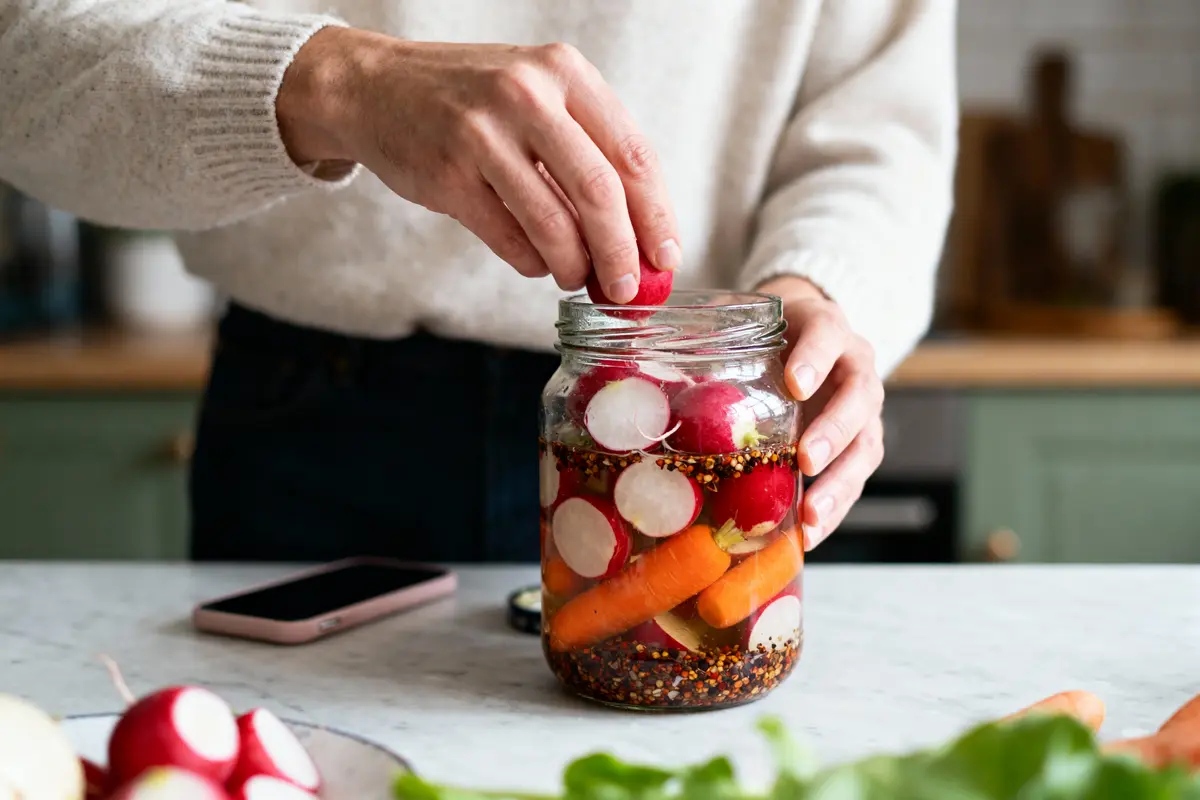 Personne mettant des radis dans un bocal de légumes marinés sur un plan de travail en cuisine.
