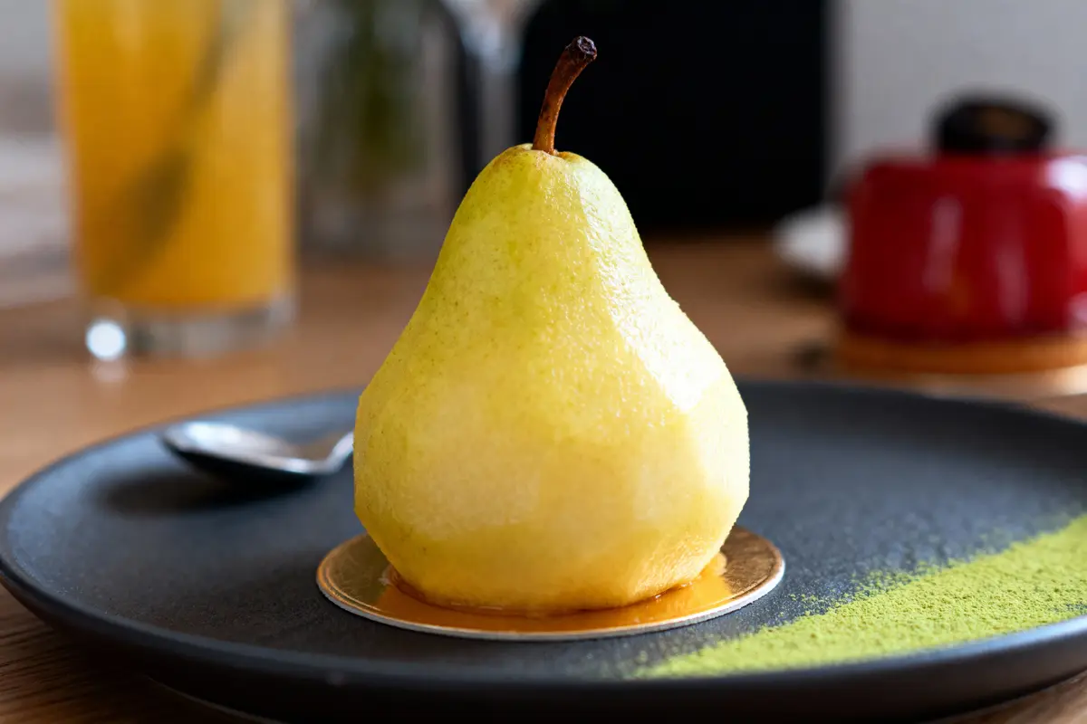 Poire entière sur assiette noire avec poudre verte, verre et objet rouge flous à l'arrière-plan.