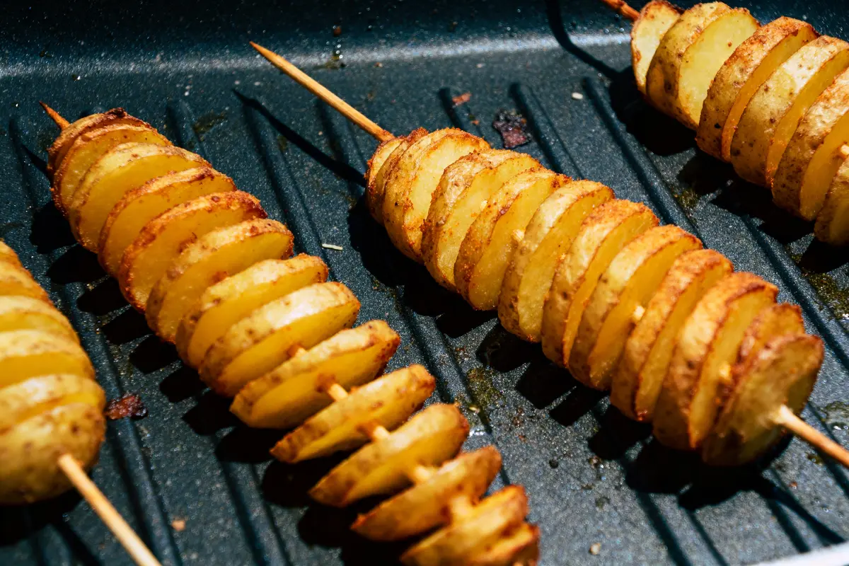 Tranches de pommes de terre en spirale sur des brochettes, grillées sur une poêle noire.