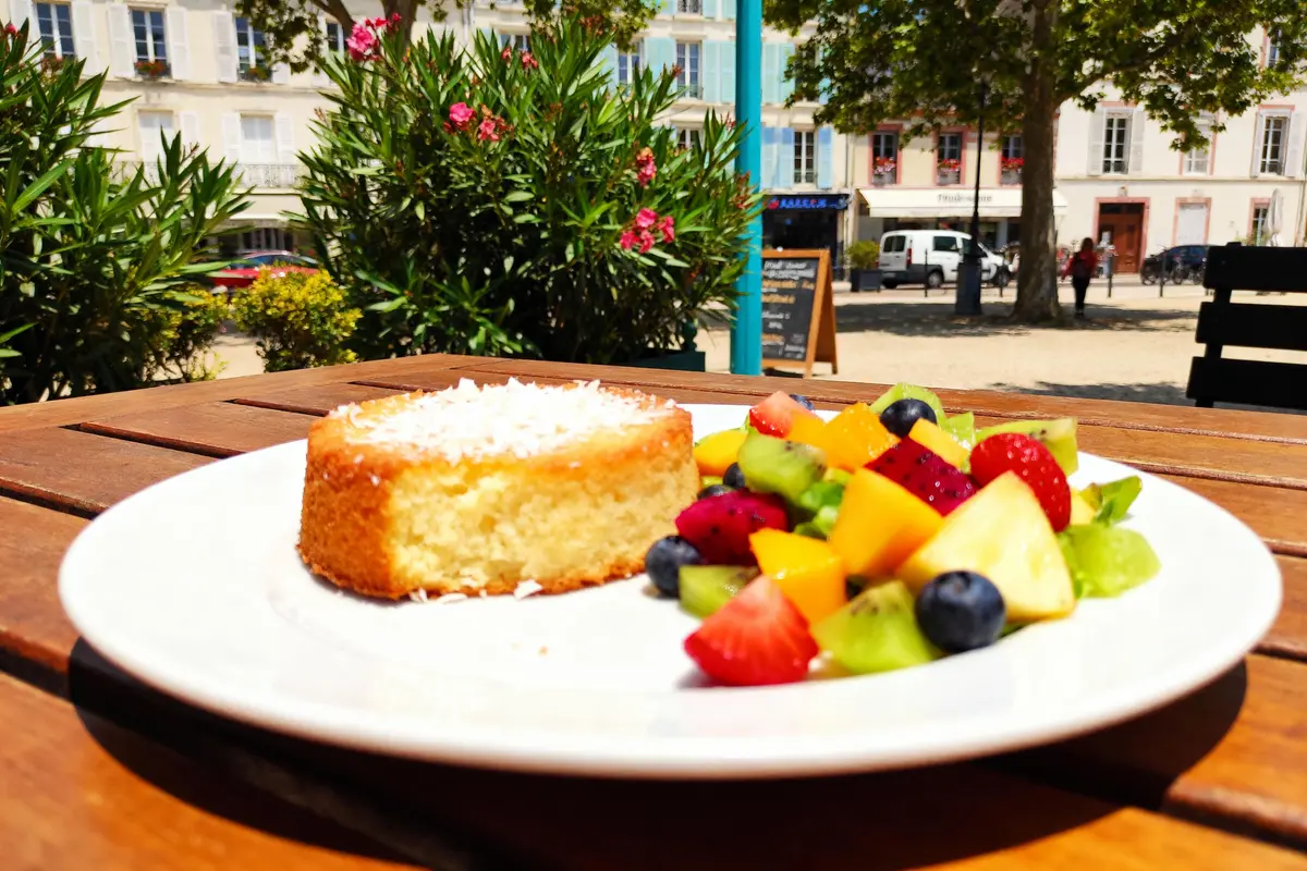 Assiette de gâteau et salade de fruits colorée sur table en terrasse avec plantes et bâtiments en arrière-plan.