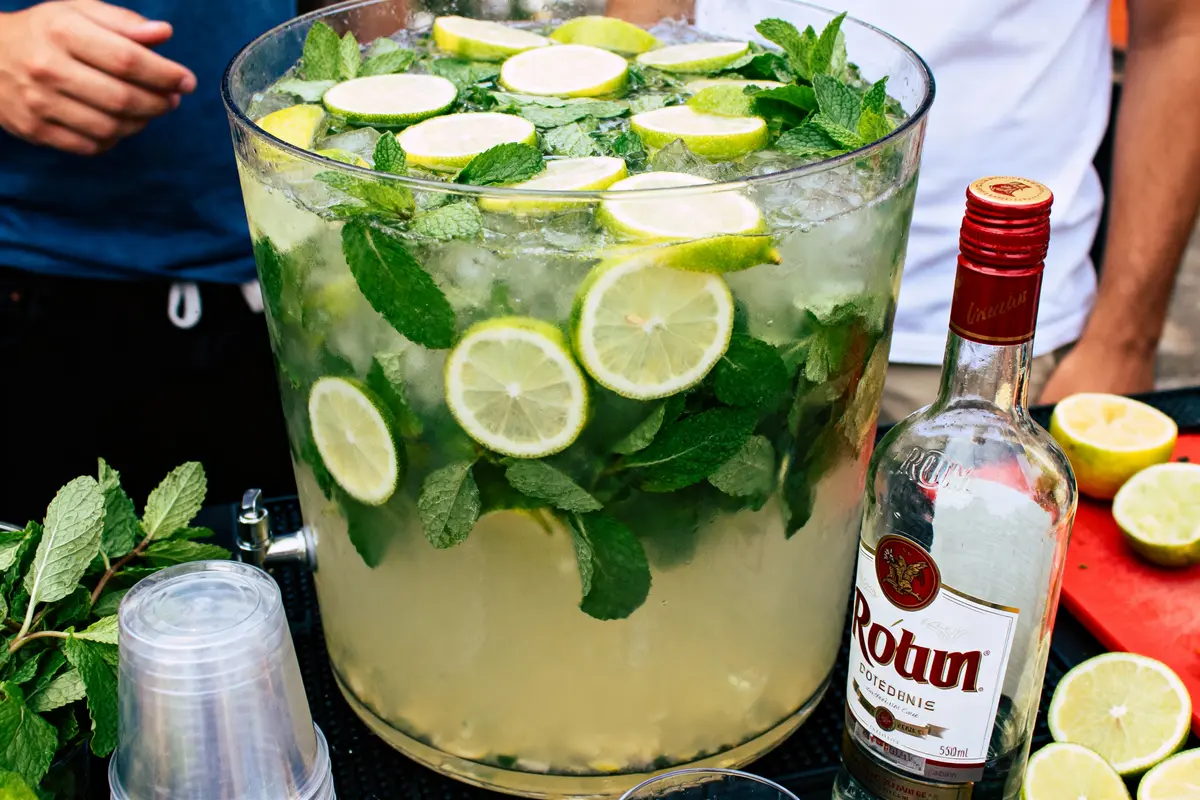 Grand récipient de mojito avec citron, menthe, bouteilles d'alcool et gobelets en plastique autour.