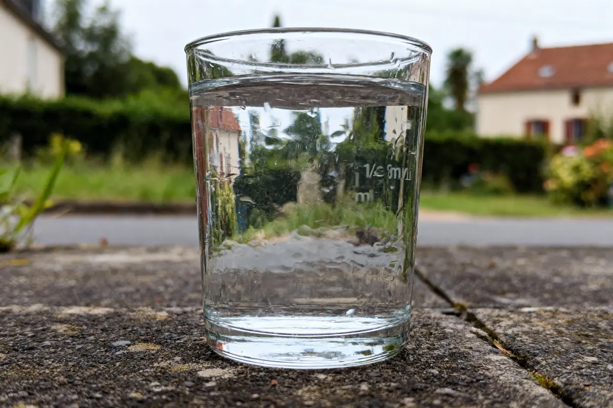 Verre d'eau posé sur une surface extérieure, reflétant un paysage flou en arrière-plan.