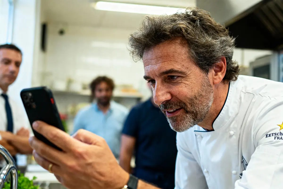 Chef en veste blanche souriant, tenant un smartphone, avec deux personnes floues en arrière-plan dans une cuisine.
