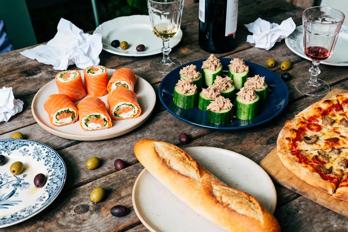 Assorted appetizers on a rustic wooden table with wine, including salmon rolls, cucumber tuna cups, pizza, and baguette.