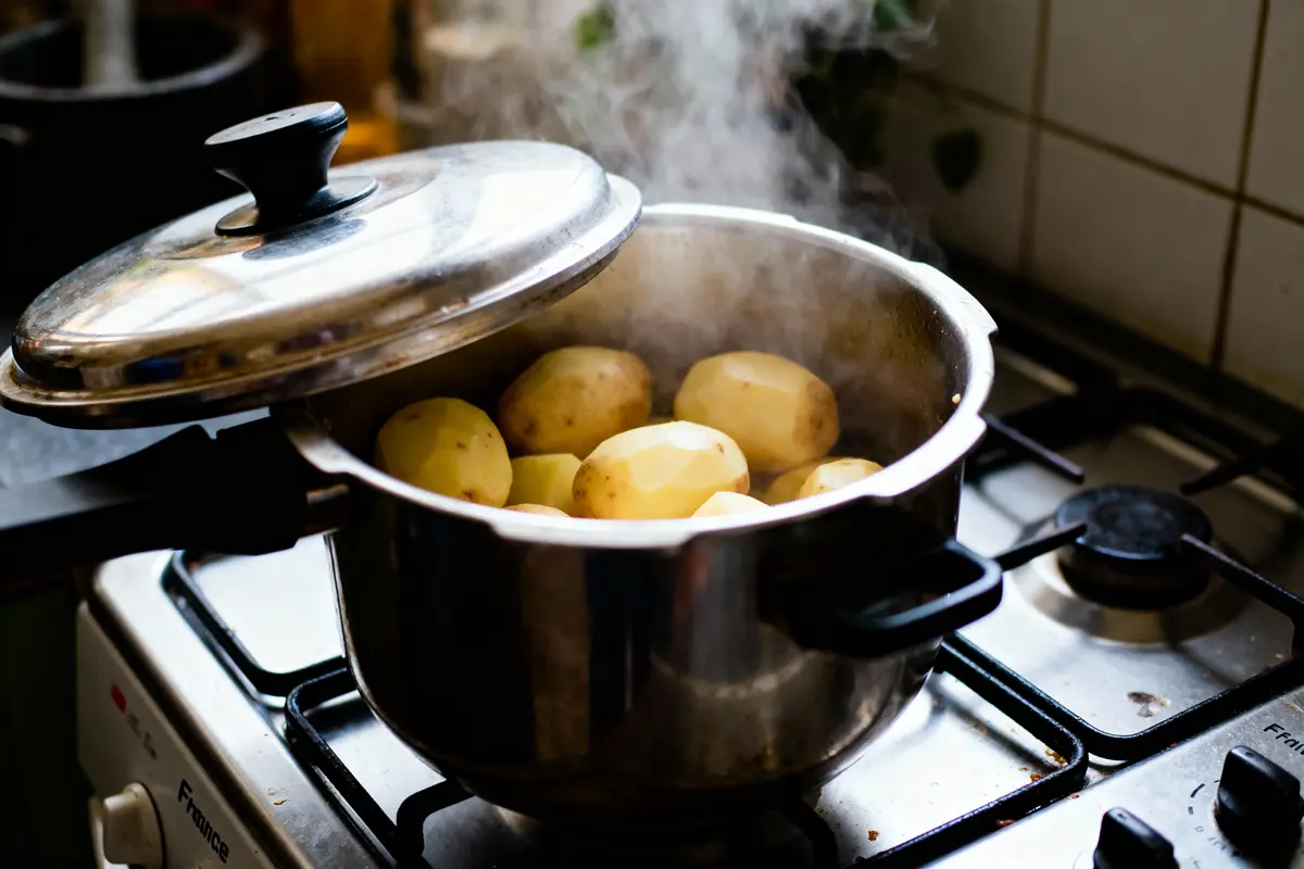Casserole en acier inoxydable contenant pommes de terre épluchées, sur une cuisinière avec couvercle partiellement fermé, vapeur visible.
