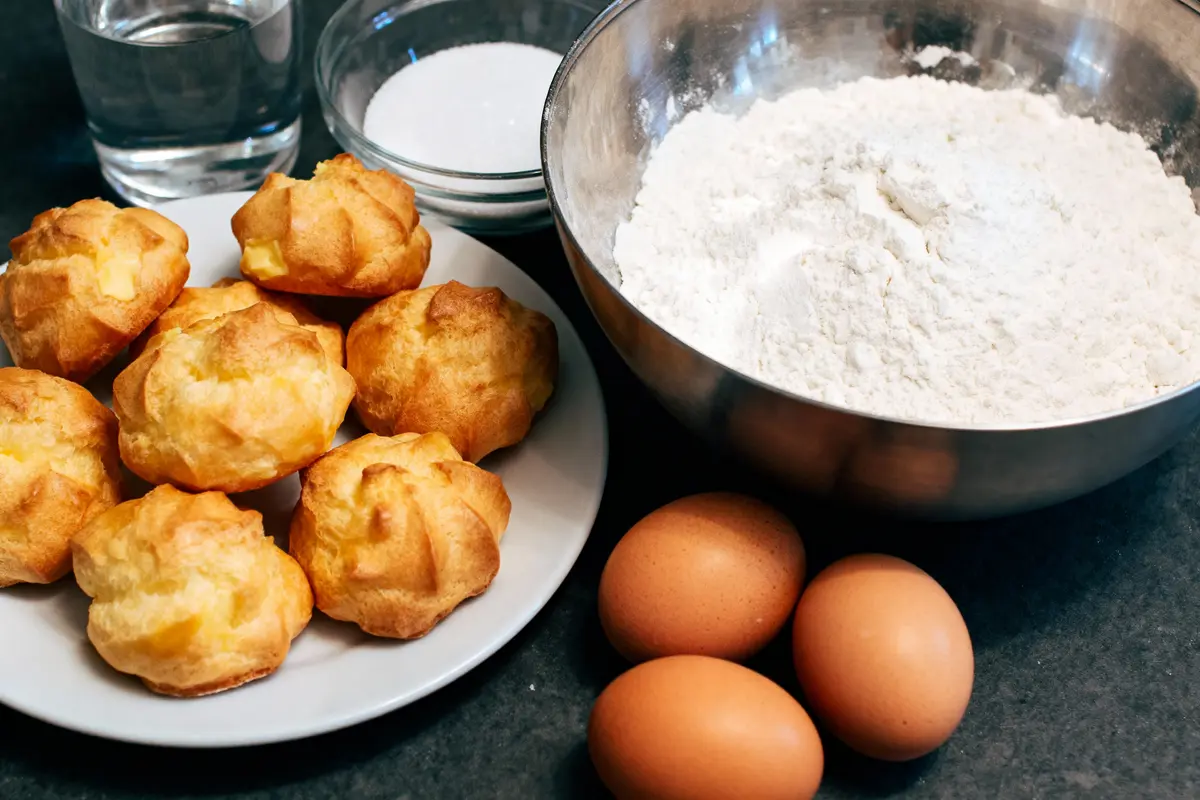 Choux pâtissiers sur assiette avec œufs, farine, sucre, et verre d'eau sur une table.