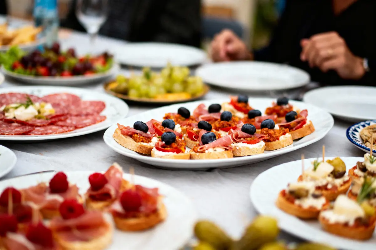 Assiette de hors-d'œuvre variés avec fruits, charcuteries et fromage sur table, personnes en arrière-plan.
