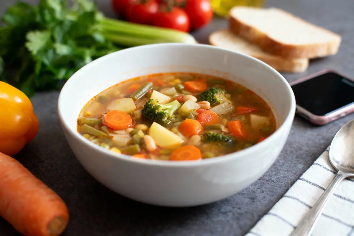 Bol de soupe aux légumes avec pain, légumes, et téléphone sur table grise.