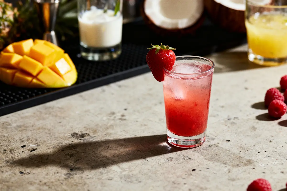 Verre de cocktail rouge avec fraise, mangue, framboises, et boissons en arrière-plan sur comptoir.
