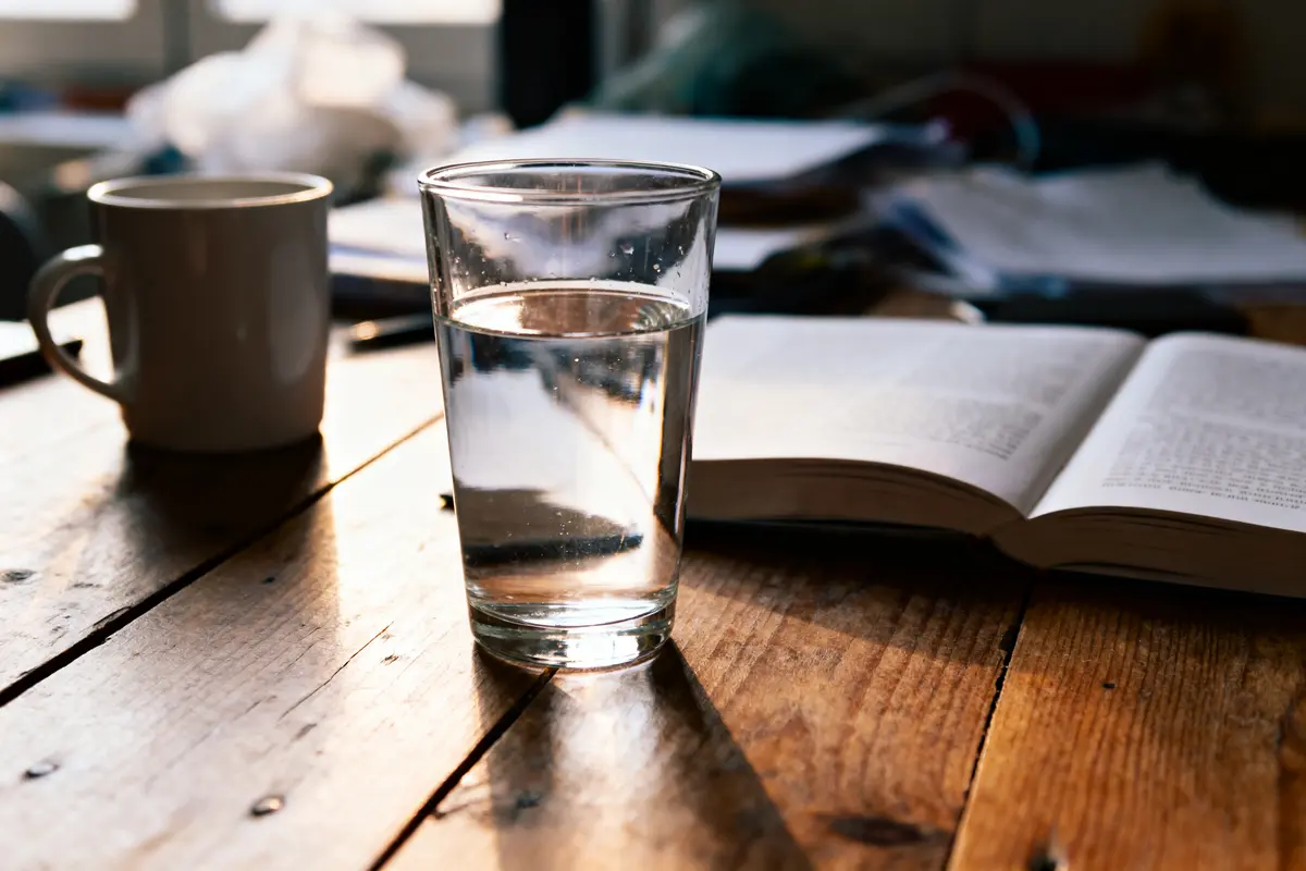 Verre d'eau, tasse, livre ouvert sur table en bois illuminée par la lumière naturelle.