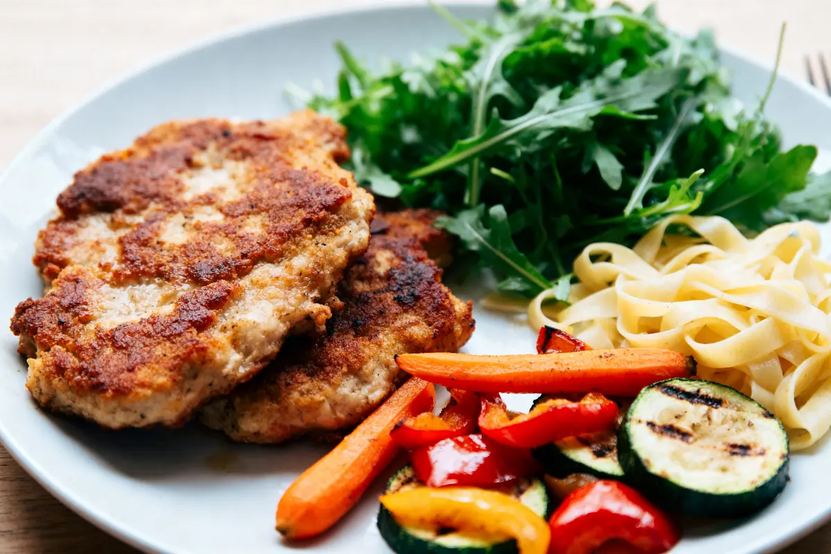 Assiette avec escalope panée, légumes grillés, roquette et tagliatelles. Plat coloré et appétissant.