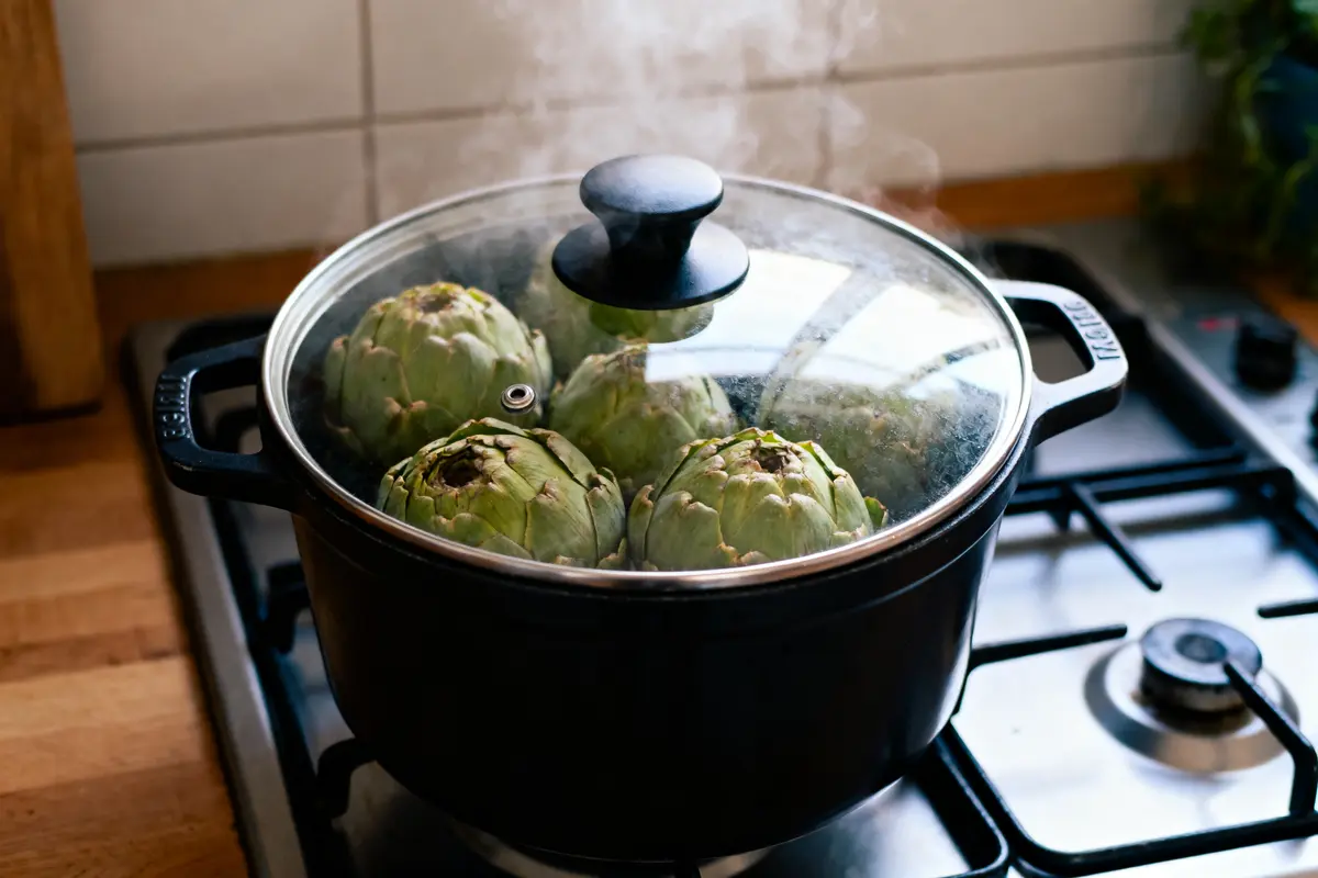 Casserole sur feu contenant quatre artichauts cuits à la vapeur sous un couvercle transparent.