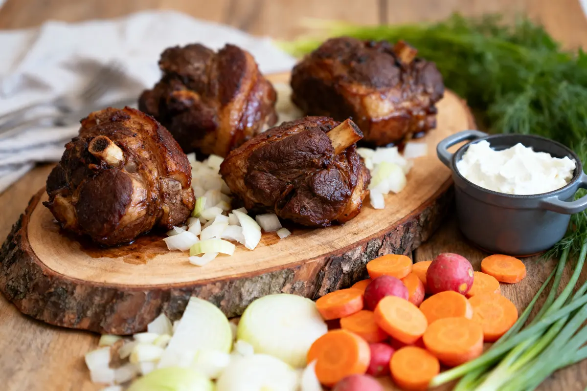 Trois jarrets de porc cuits sur plateau bois, accompagnés d'oignons, carottes, radis, crème et herbes fraîches.