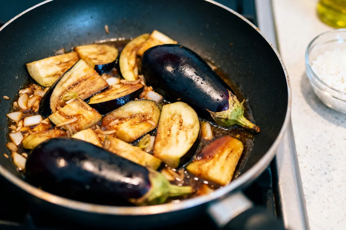 Aubergines en tranches et entières cuisant dans poêle avec oignons bruns, plan de travail cuisine proche.