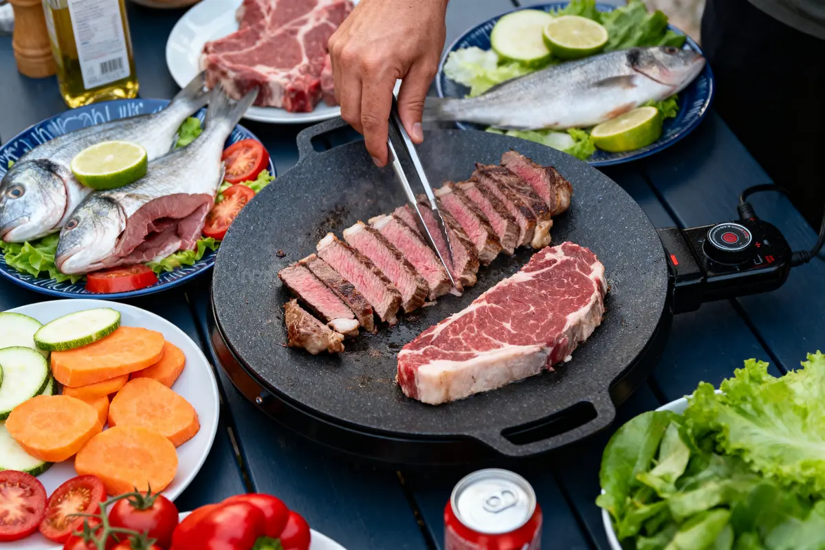 Barbecue avec viande, poisson, légumes, main utilisant des pinces, surface de cuisson et boissons en arrière-plan.