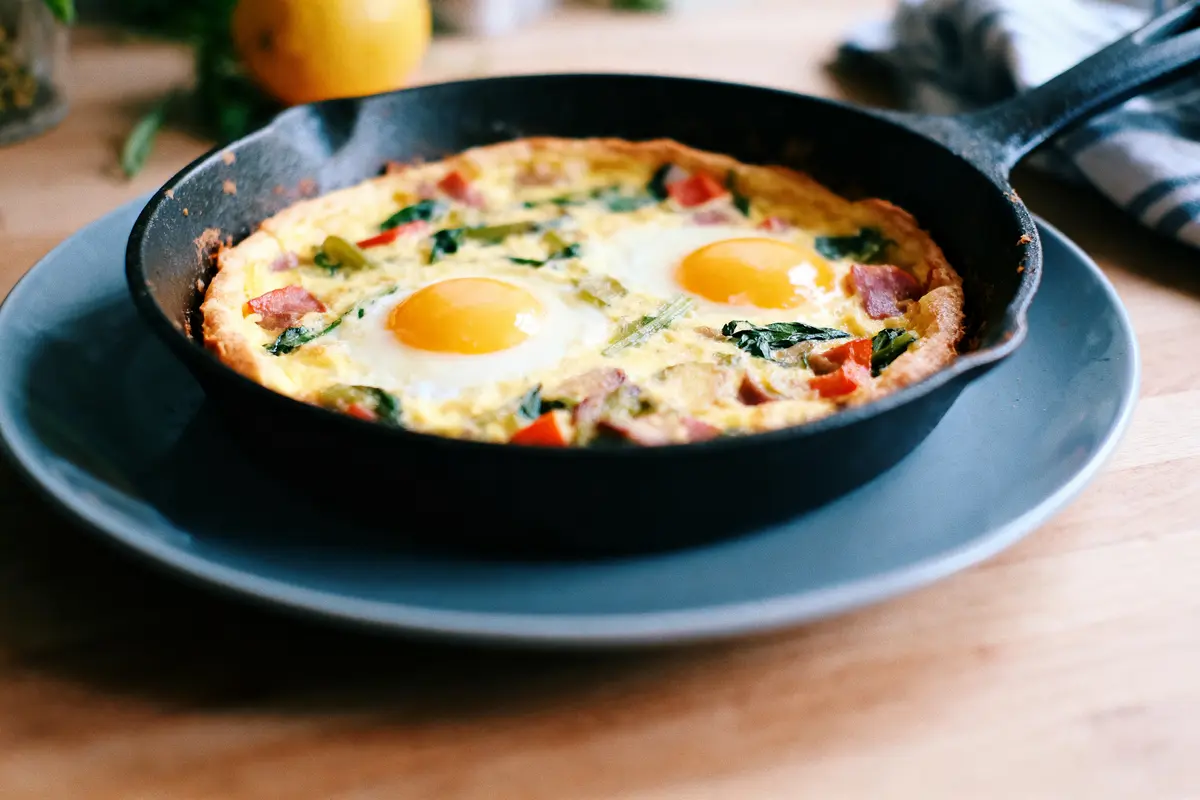 Omelette avec légumes et deux œufs au plat dans une poêle en fonte sur une assiette grise.