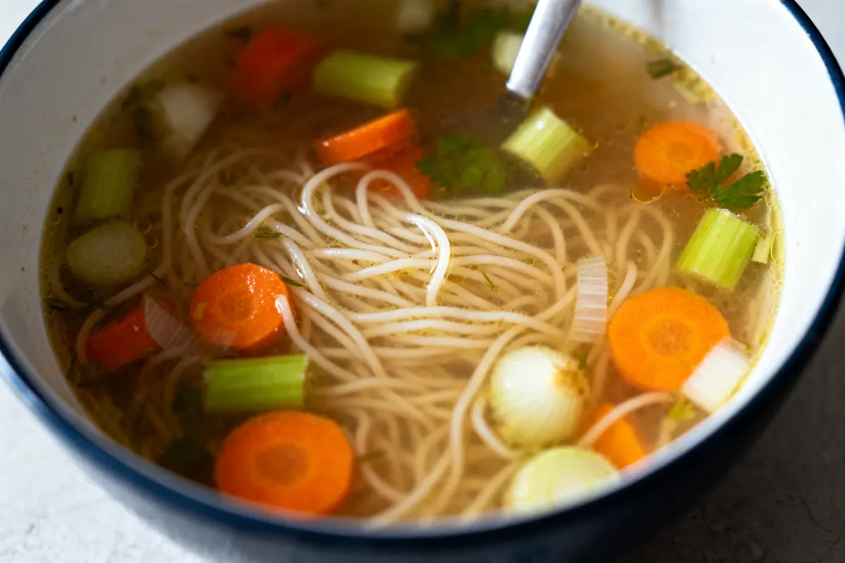 Bol de soupe avec des nouilles, carottes, céleri, oignons et herbes, cuillère en métal visible.